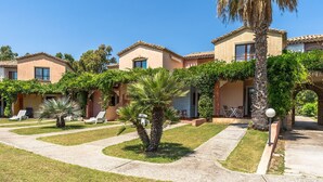 A row of two-story buildings with a garden in front, featuring palm trees and a pathway. - One bedroom apartment (4 people) Premium - Residence Baia delle Palme (Sardegna)