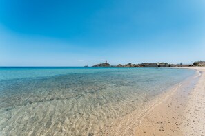 A clear beach with calm waters, a sandy shore, and a rocky outcrop in the distance. - One bedroom apartment (4 people) Premium - Residence Baia delle Palme (Sardegna)