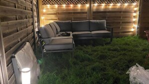 Une clôture en bois décorée de guirlandes lumineuses, un canapé d’angle gris et une petite table.