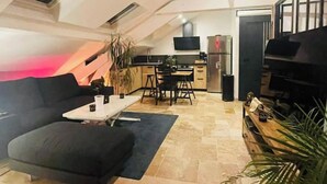 Un salon moderne avec un canapé noir, une petite table basse et un téléviseur. Il y a également un coin cuisine équipé d’un réfrigérateur, d’un four à micro-ondes et d’une table à manger.