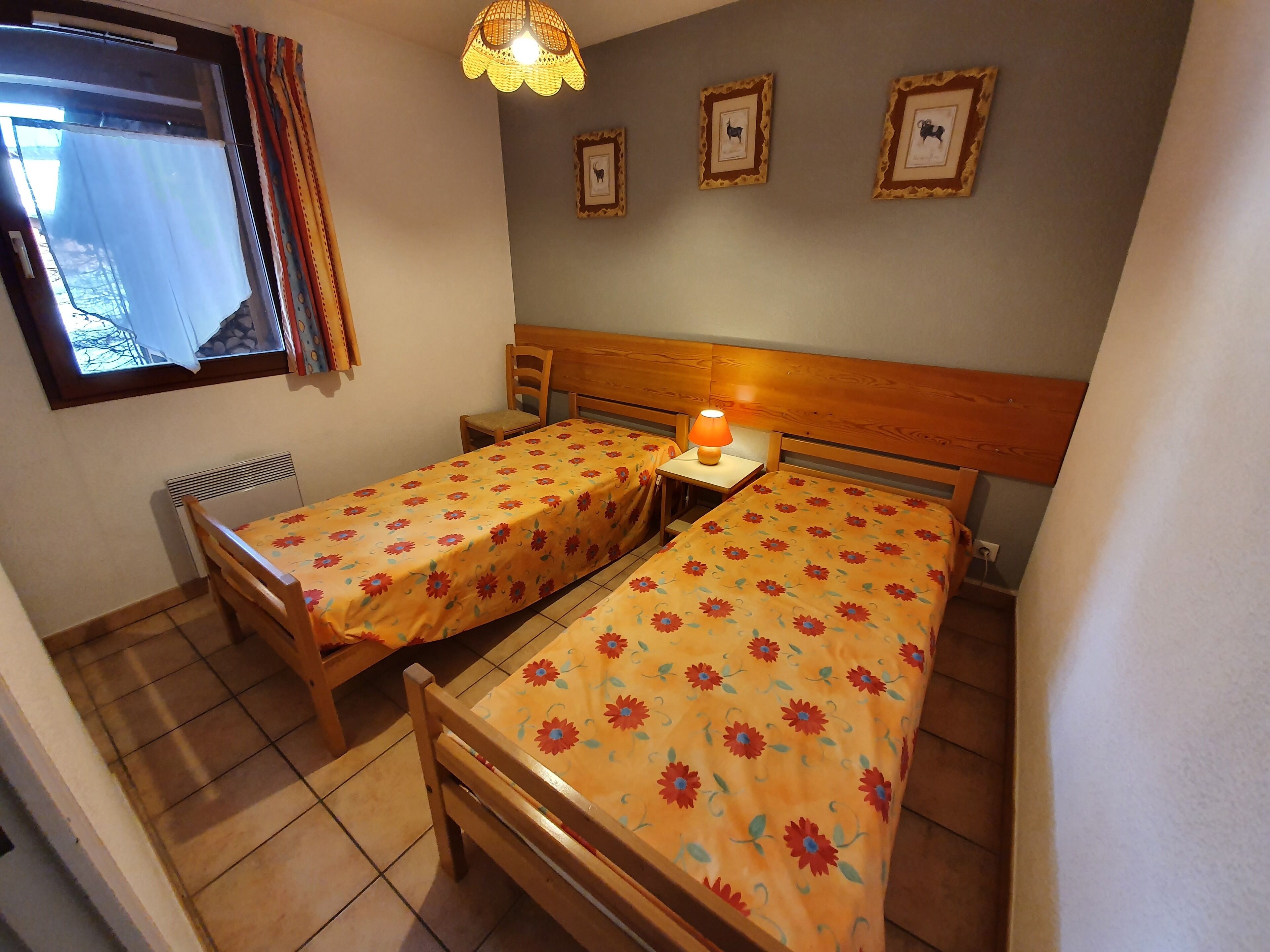Une chambre avec deux lits en bois, recouverts de couvre-lits à motifs floraux, une petite table avec une lampe, une chaise, un radiateur et une fenêtre avec des rideaux.