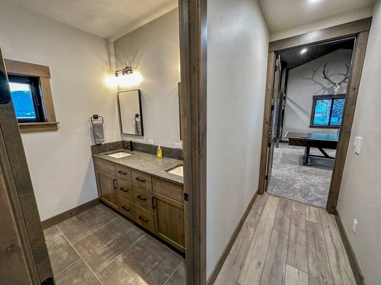 A modern bathroom with a vanity, sink, and mirror. A window provides natural light, and a door leads to a room with a ping pong table.