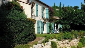 Une villa traditionnelle de style méditerranéen, dotée d’un escalier en pierre menant à une porte en bois, et entourée d’une végétation luxuriante et d’un mur en pierre.