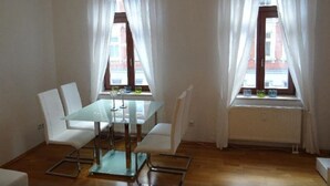 A room with a glass-top dining table, white chairs, and two windows with white curtains.
