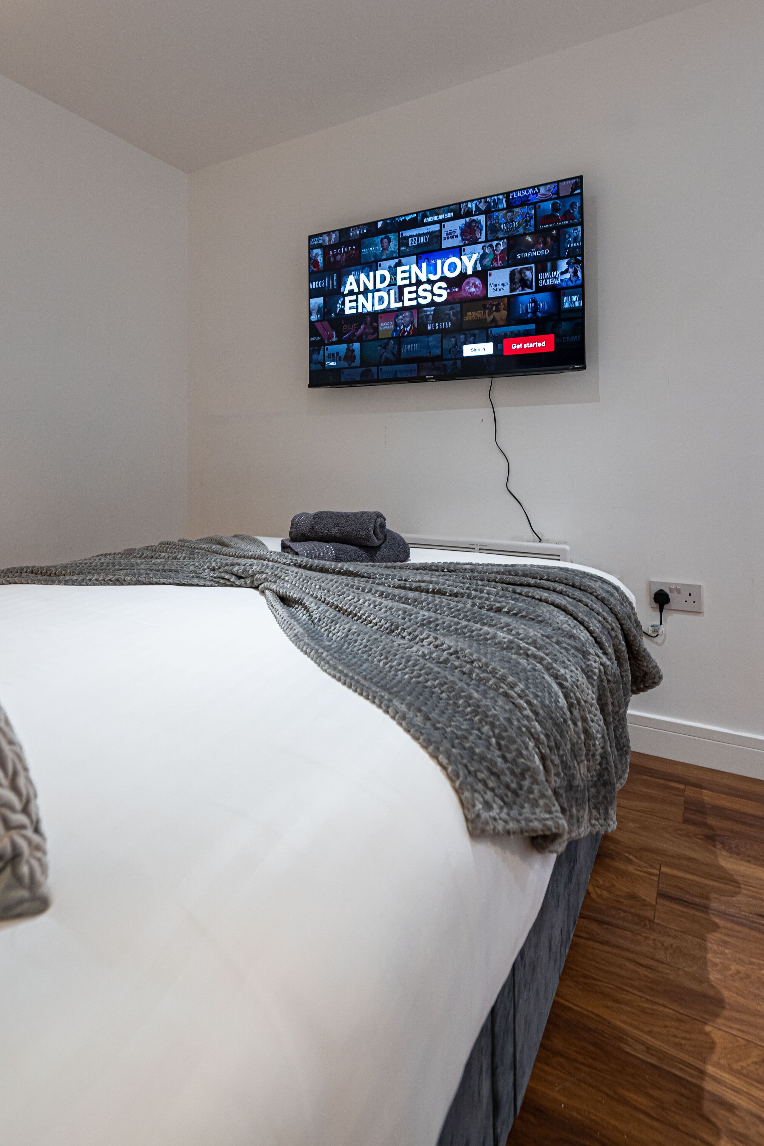 A bedroom with a bed, a TV mounted on the wall displaying a streaming service, and a power outlet on the wall.
