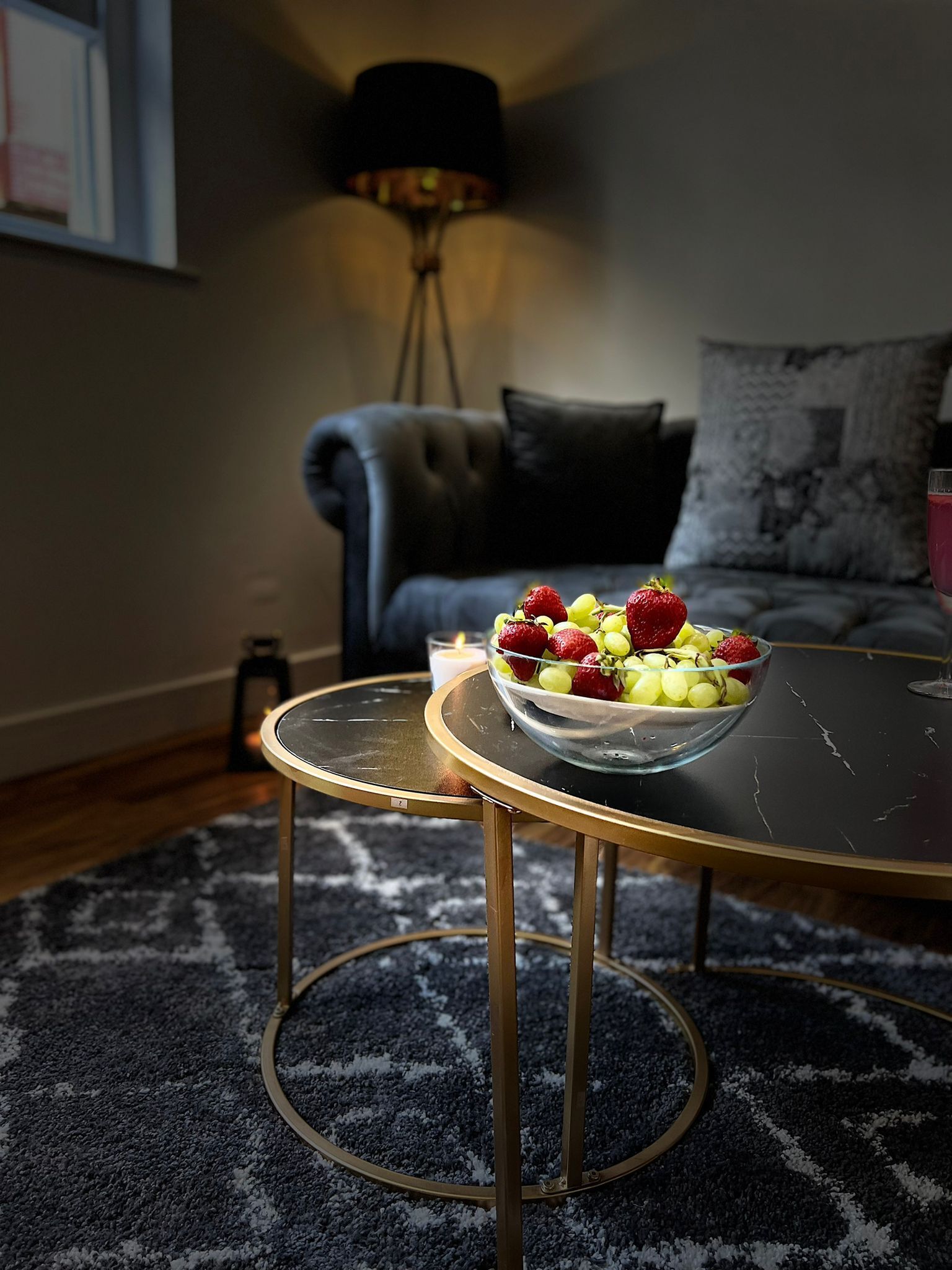 Een ronde glazen tafel met metalen poten, een schaal vol gemengd fruit en een kaars op een kamerkleed met een patroon in een woonkamer met een donkere bank en een lamp op de achtergrond.