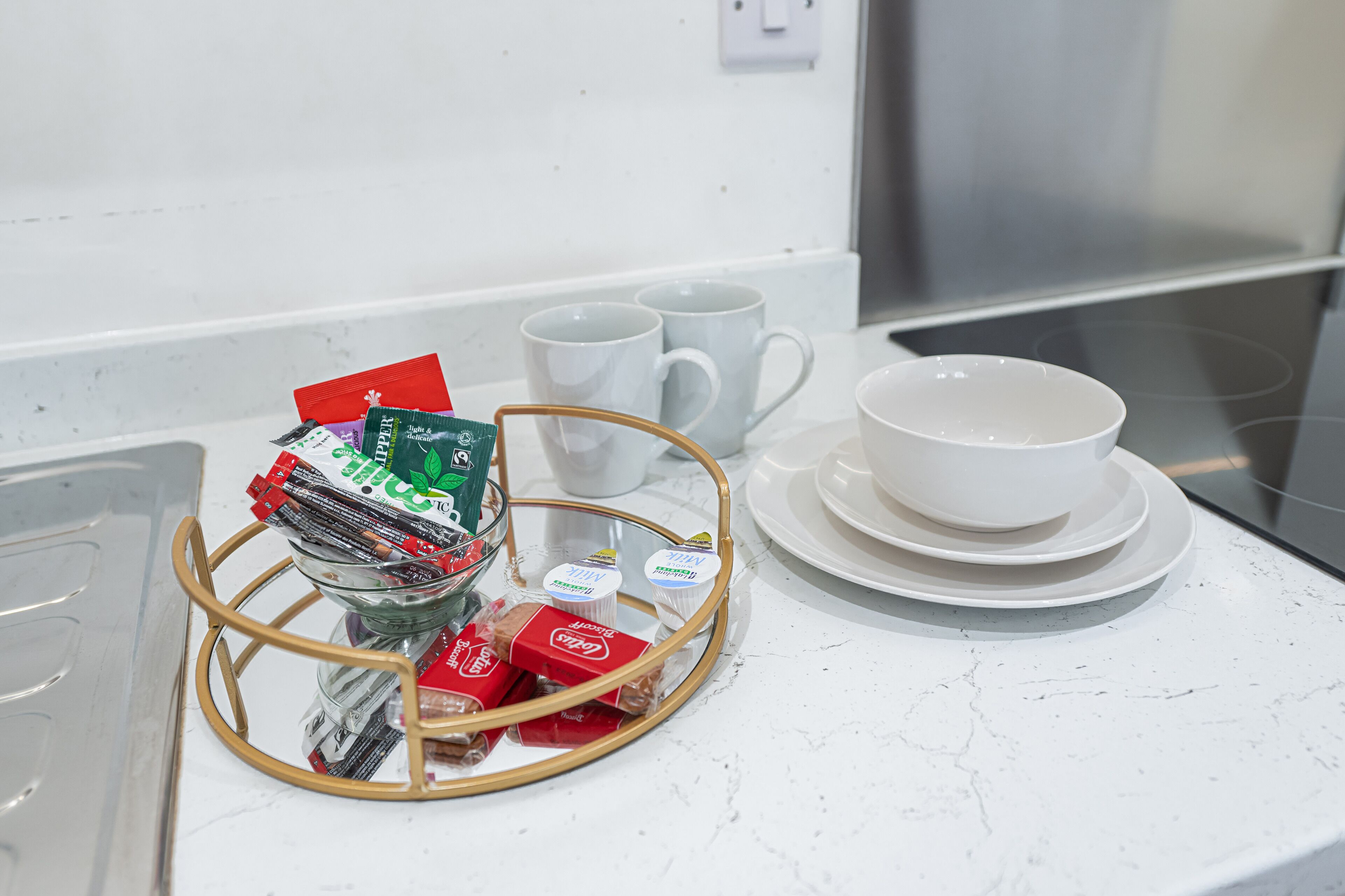A kitchen counter with a white marble surface, a metal tray with packaged food items, two white mugs, and stacked white plates.