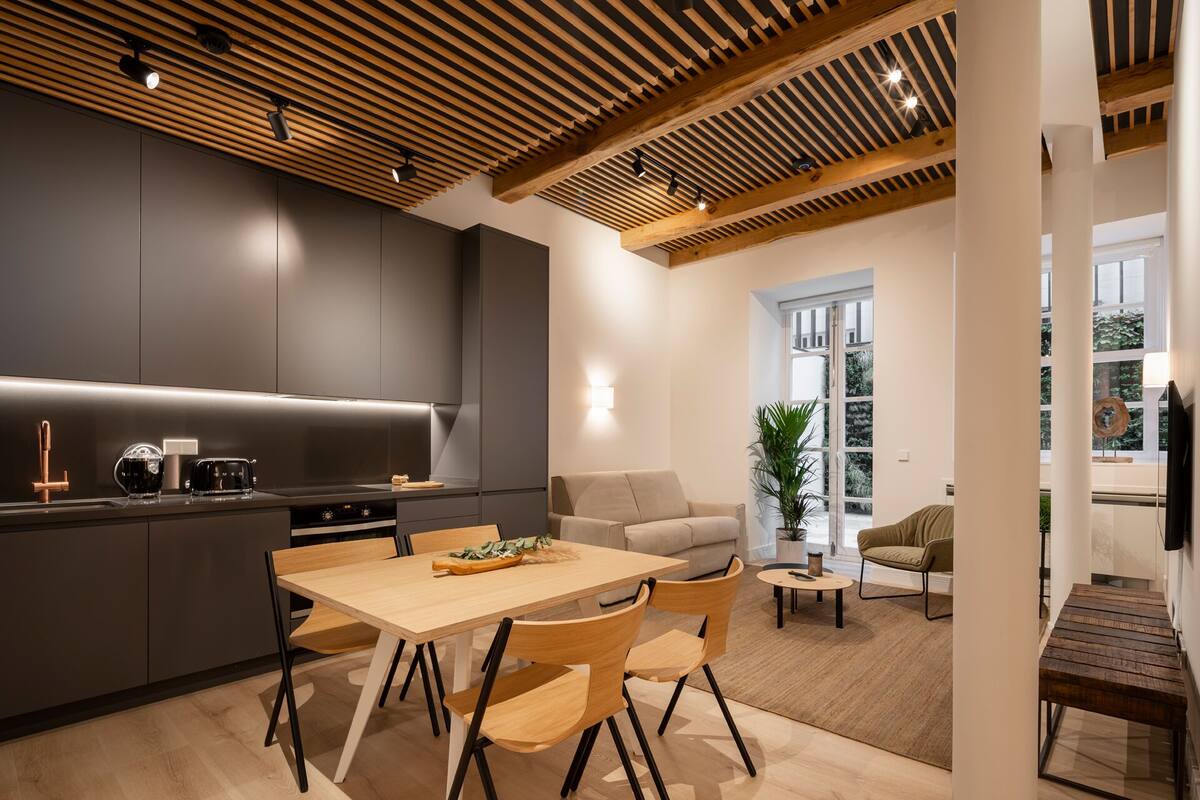 A modern kitchen with a wooden ceiling, a dining area with a table and chairs, and a living space with a sofa and a plant.
