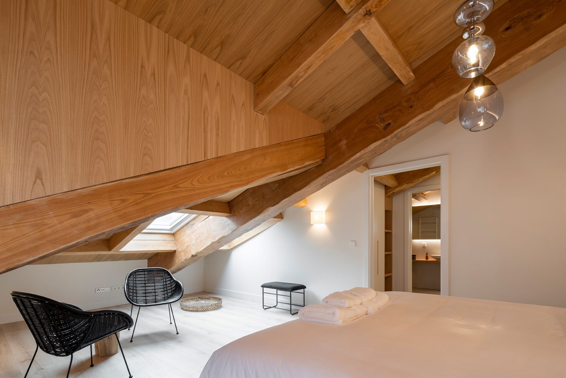 A bedroom with a sloped ceiling, wooden beams, a bed with white linens, a black chair, a small stool, and a skylight.