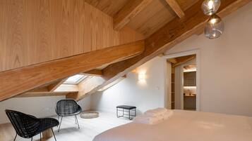 A bedroom with a sloped ceiling, wooden beams, a bed with white linens, a black chair, a small stool, and a skylight.