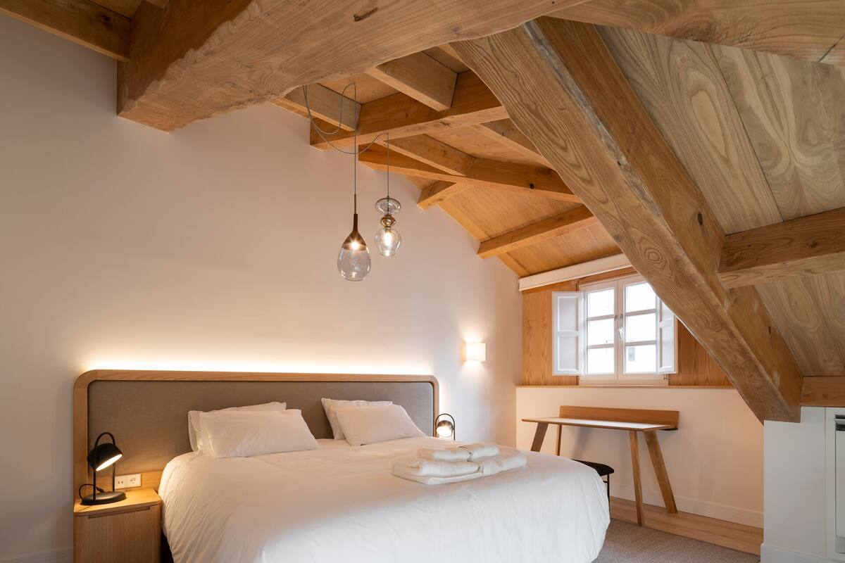 A bedroom with a wooden ceiling, a bed with white linens, a bedside table with a lamp, and a window with natural light.