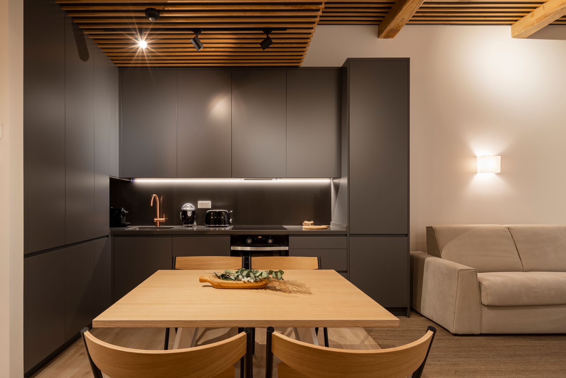 A modern kitchen with dark cabinetry, a wooden ceiling, and a dining area with a table and chairs.