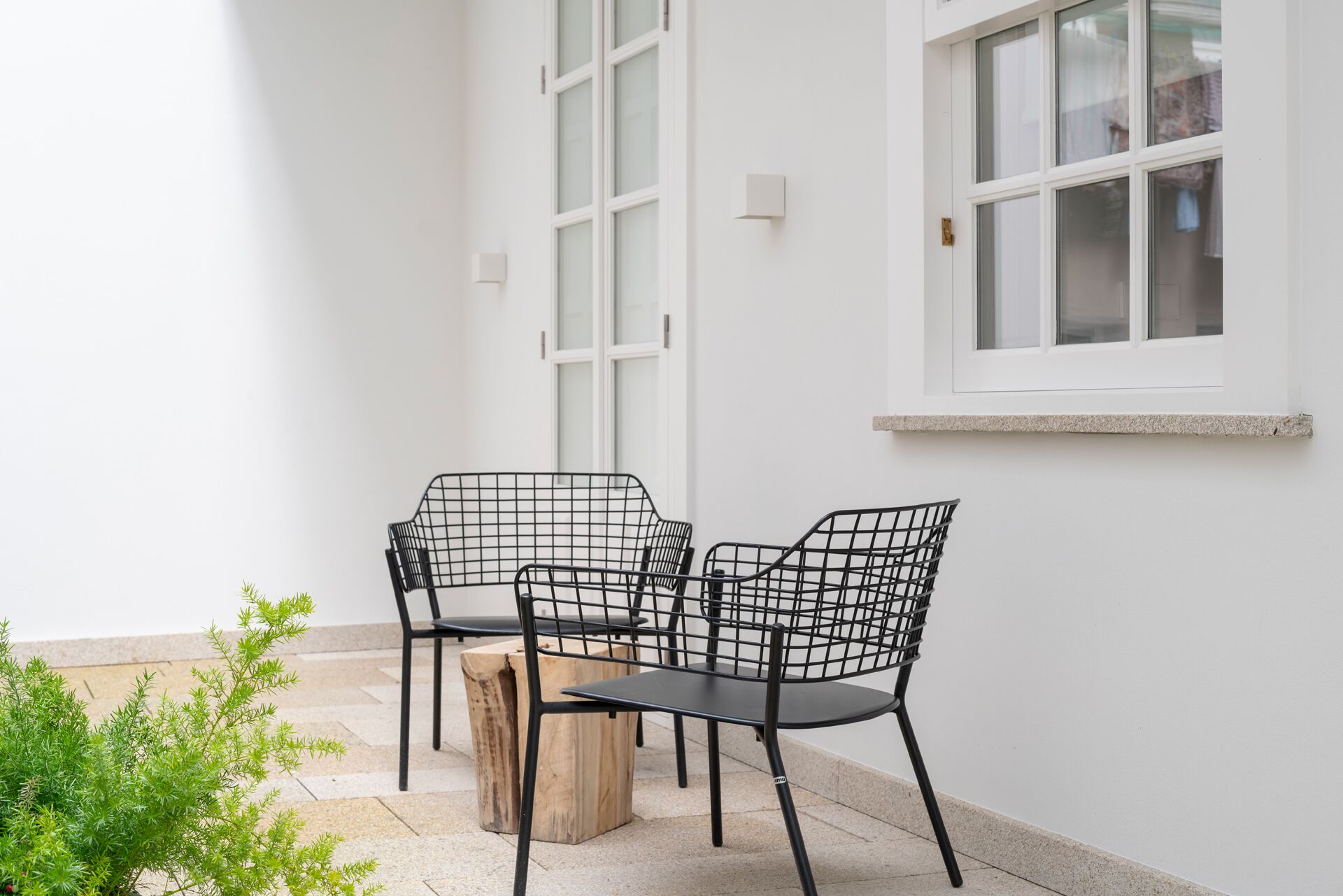 A patio area with two black wire mesh chairs and a table, a wooden stump, and a small green plant.