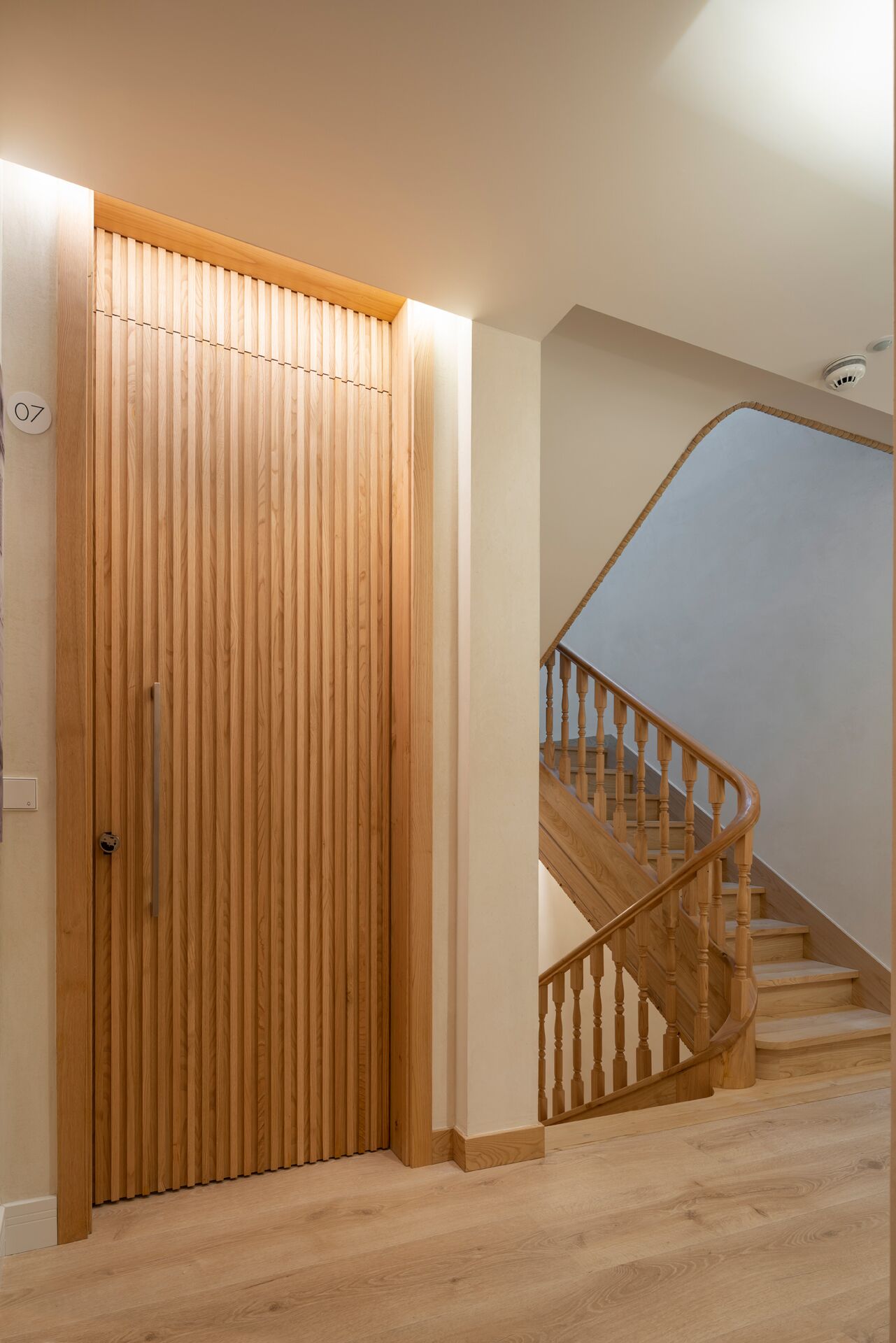 Una puerta de madera con un picaporte vertical, una escalera de madera con barandilla y un interruptor de luz en la pared.