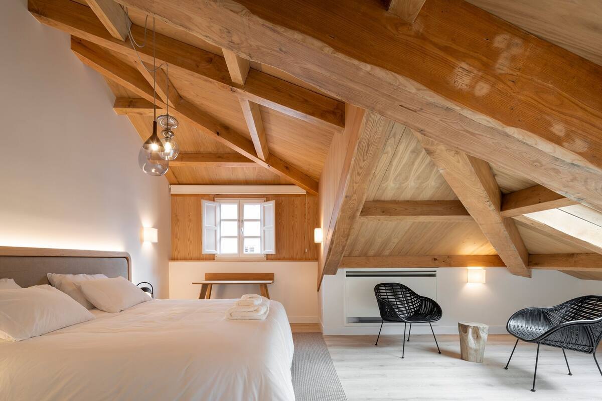 A bedroom with a large bed, a wooden ceiling, a window, and two black chairs.