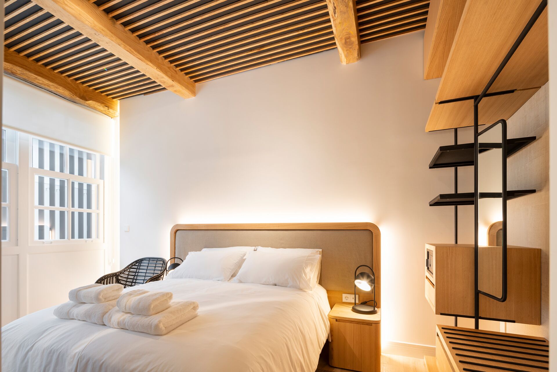 Habitación de hotel con cama, mesita de noche, lámpara y vistas al exterior.