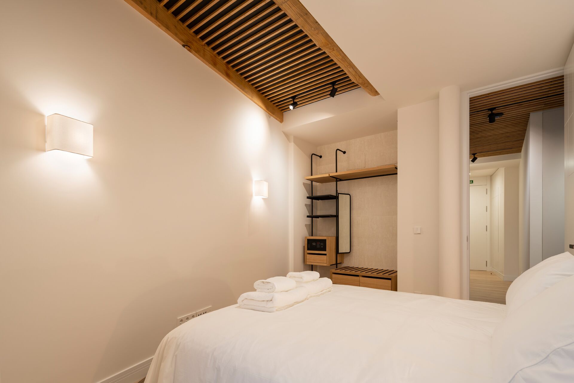 A bedroom with a bed, a bedside table with towels, a wardrobe with a mirror, and a ceiling with wooden beams.