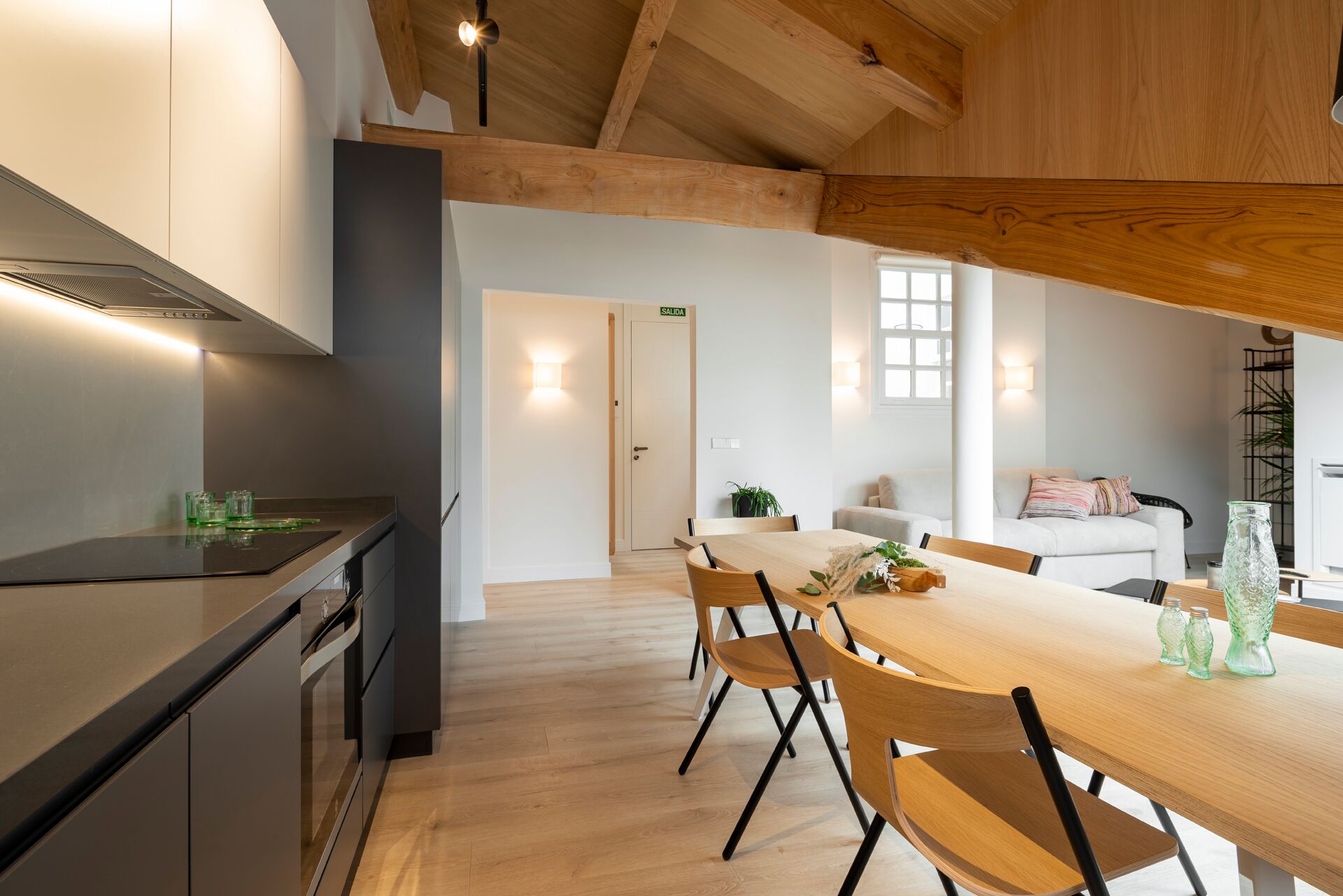 A modern kitchen with a wooden ceiling, a dining area with a wooden table and chairs, and a white sofa in the background.