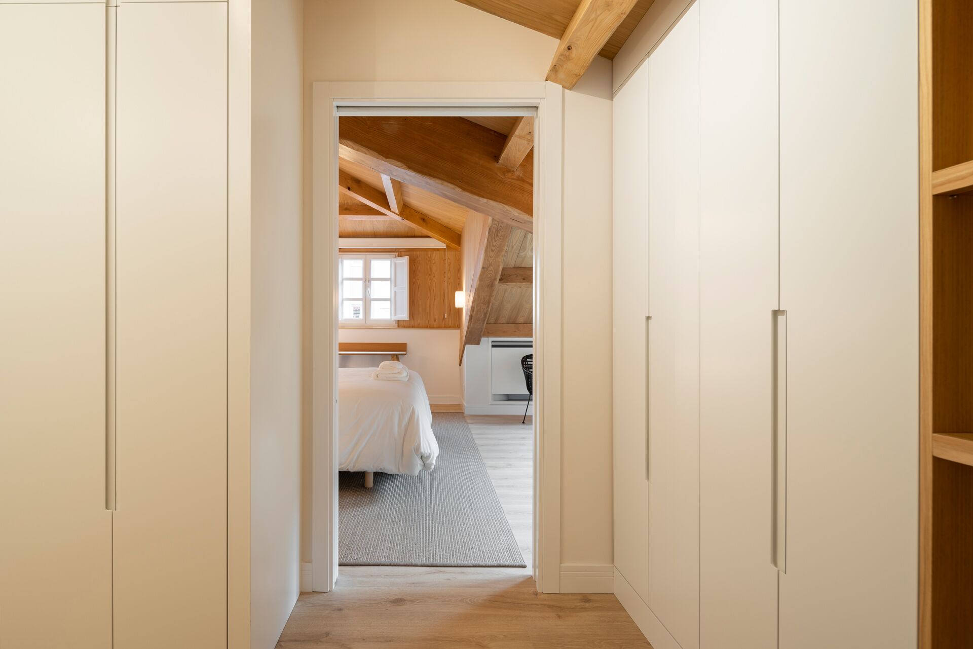 A narrow hallway leading to a bedroom with a sloped ceiling, a bed, and a window.