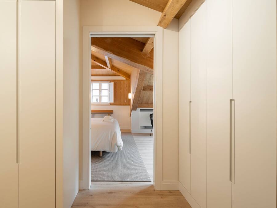 A narrow hallway leading to a bedroom with a sloped ceiling, a bed, and a window.