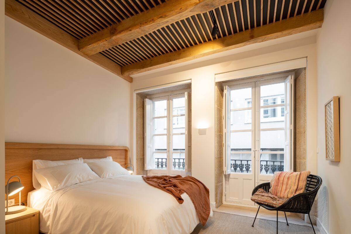 A bedroom with a wooden ceiling, a bed with white linens, a chair with a patterned throw, and a window with a view of buildings.