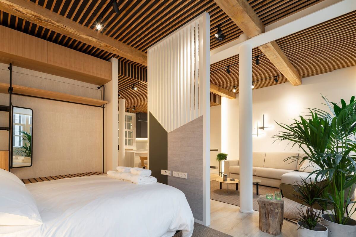 A modern hotel room with a large bed, wooden ceiling beams, a seating area with a sofa and plants, and a visible bathroom.