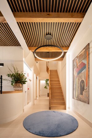 Un interior moderno con una escalera de madera, un tapete azul redondo y un techo de listones de madera con una lámpara circular.