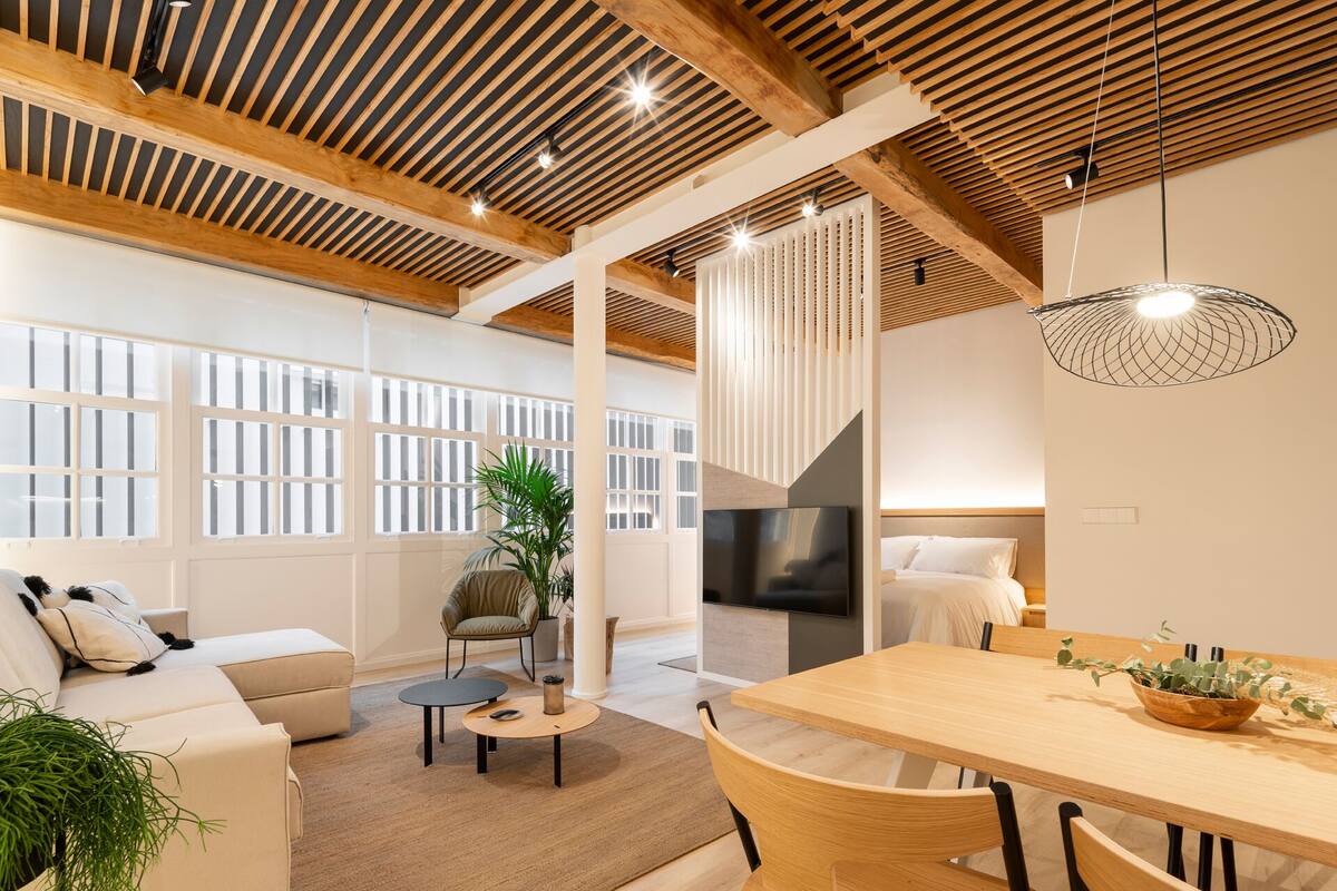 A modern living room with a wooden ceiling, large windows, a sofa, a dining table, and a TV.
