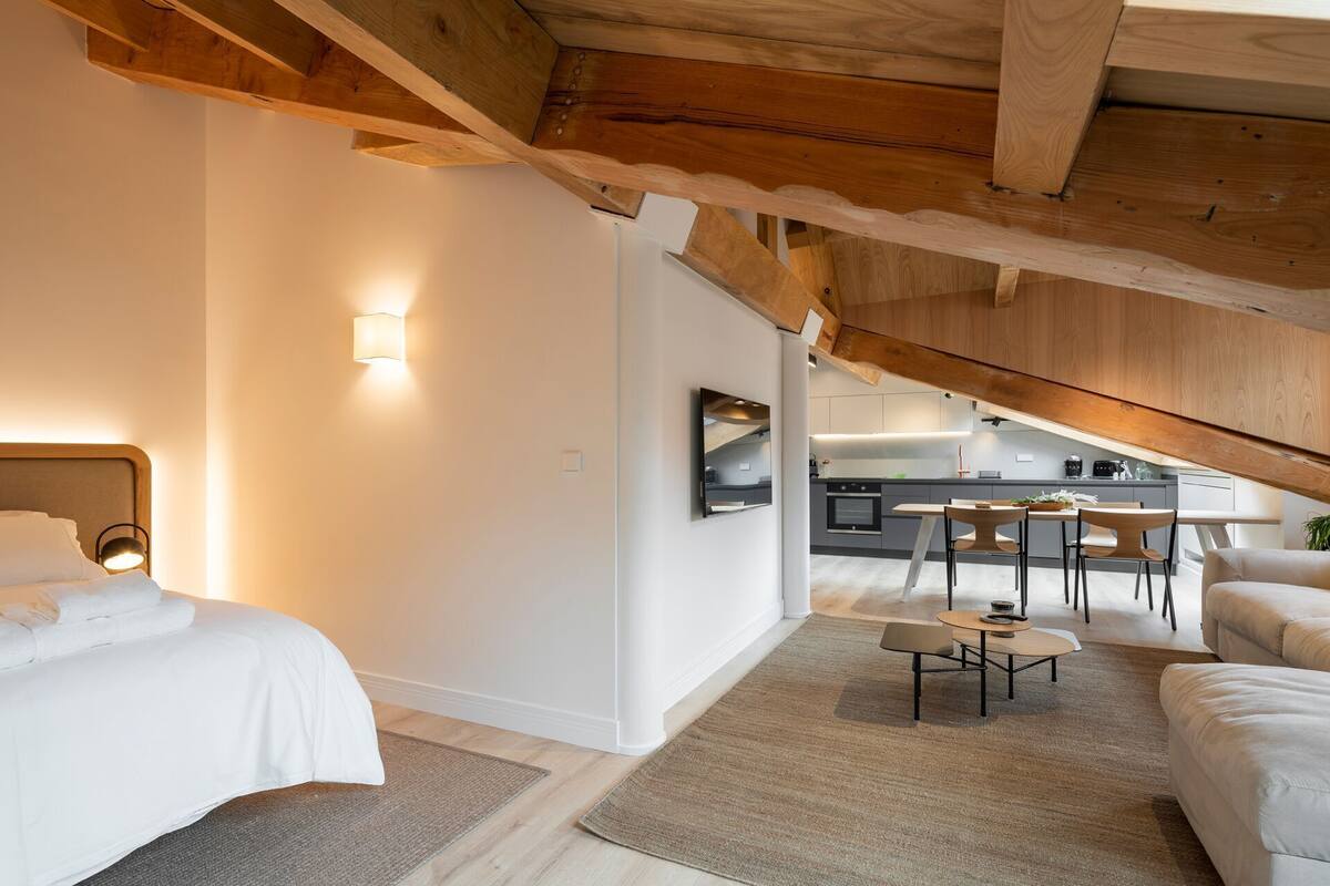 A modern loft bedroom with a large bed, a desk, and a kitchenette in the background.