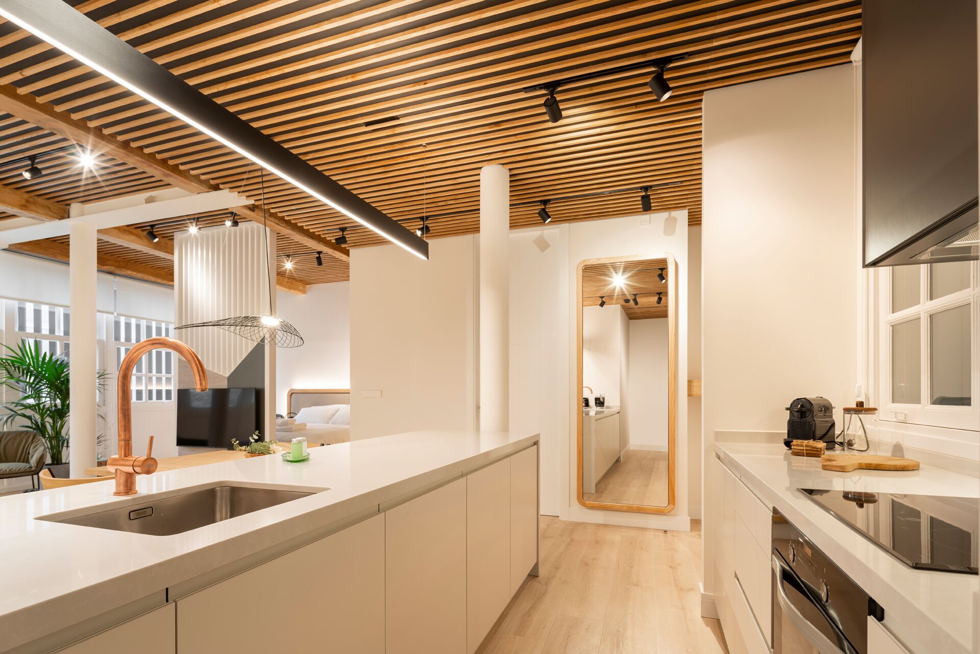 A modern kitchen with wooden ceiling beams, a large island with a sink, and a view into a bedroom.