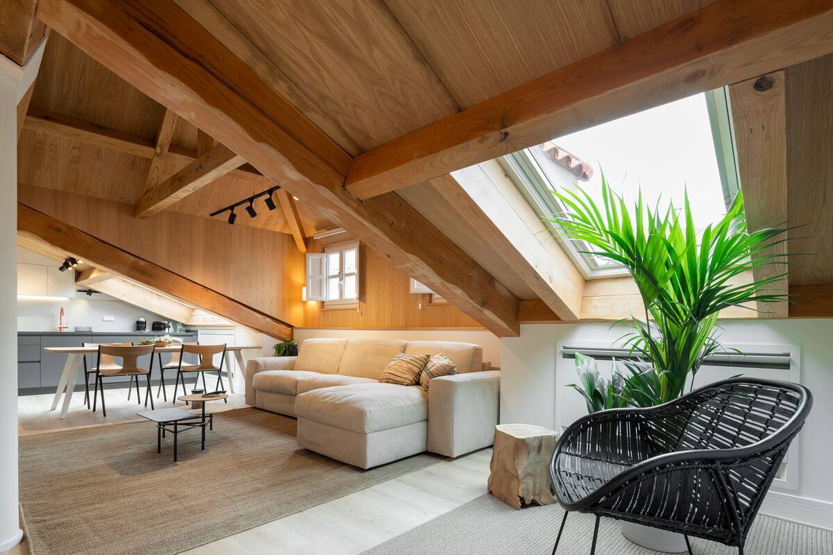 A cozy loft-style room with a high ceiling, exposed wooden beams, a dining area, a sofa, a black woven chair, and a large green plant.