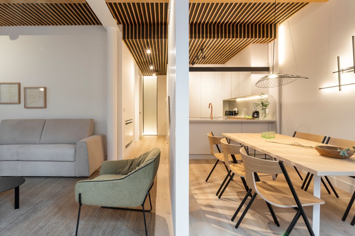 Un salon moderne avec un canapé et un fauteuil, un coin repas avec une table et des chaises en bois, et une cuisine dotée d’un comptoir et de luminaires suspendus.