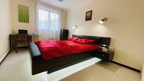 Een slaapkamer met een bed, een bureau, een stoel, een nachtkastje, een lamp en een schilderij aan de muur.