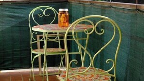 Een terras met twee groene metalen stoelen en een tafel, een potje op de tafel, en uitzicht op een bebost heuvel.'