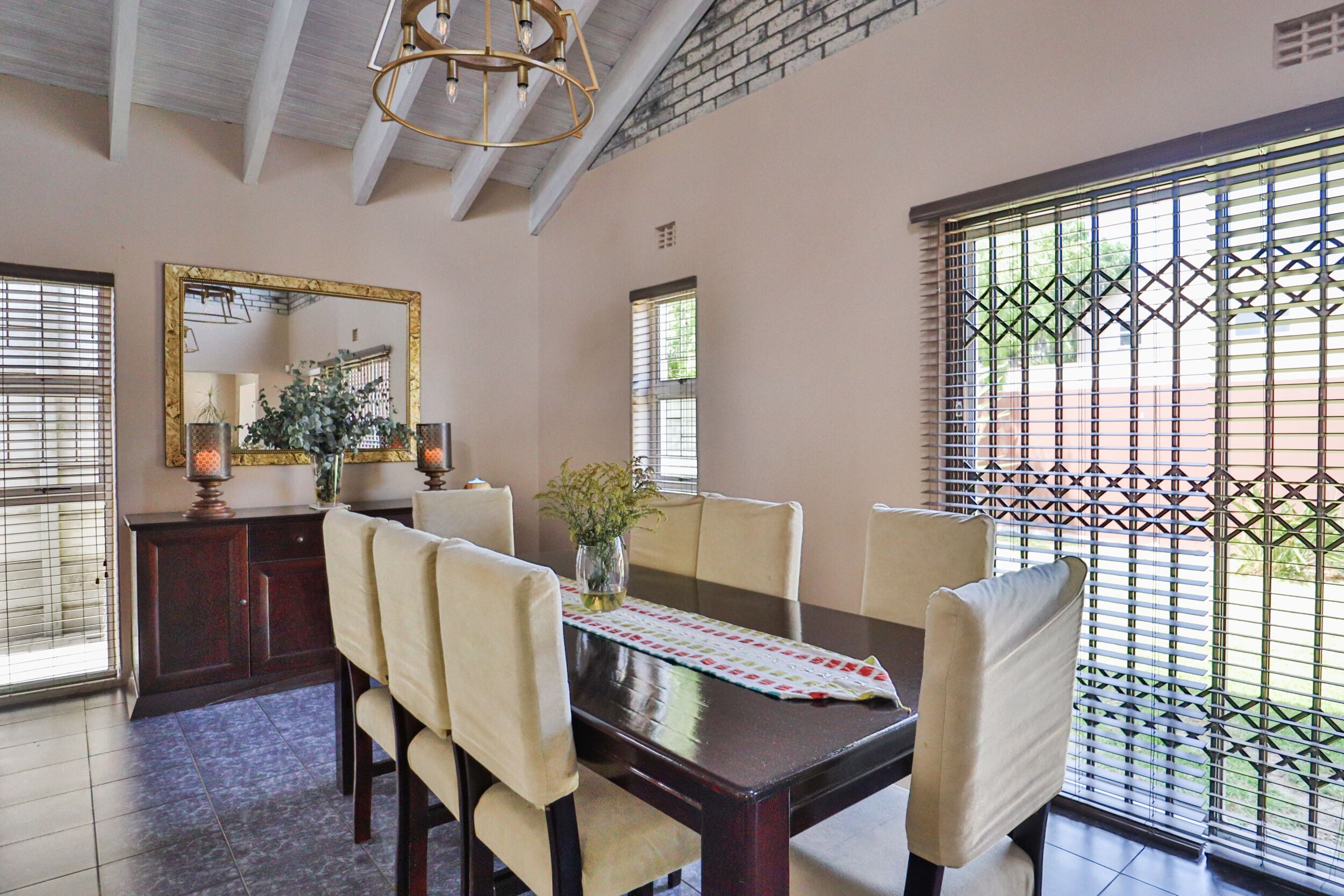 Un comedor con una mesa de madera, sillas color beige, un jarrón con flores y un espejo en la pared.