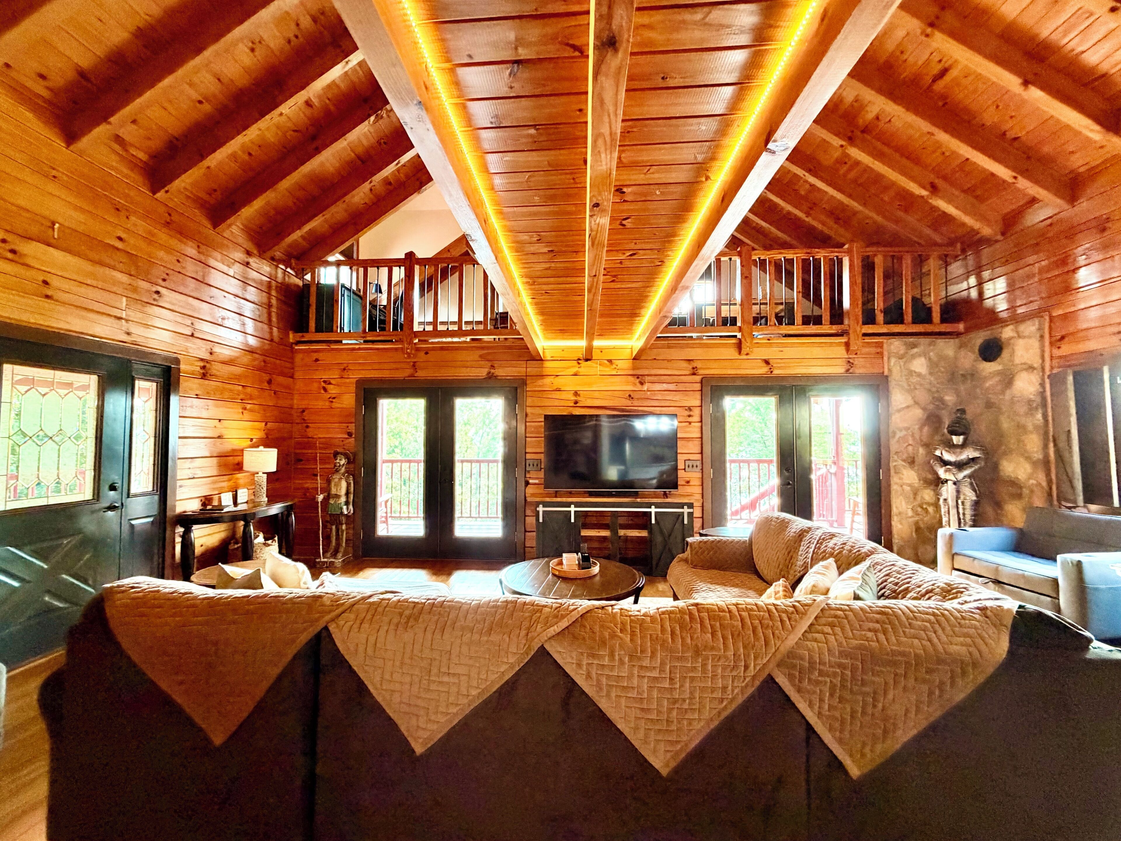 A cozy cabin interior with a loft bedroom, a living area with a sofa and a TV, and a dining table.