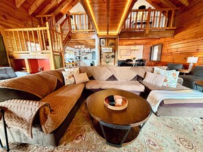 A cozy cabin interior with a large L-shaped sectional sofa, a round wooden coffee table, and a staircase leading to an upper level. - Cozy Christmas Log Cabin on Lake Eufaula/Private Dock & Fire Pit (Georgetown)