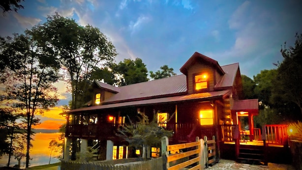 A wooden cabin with a porch, a stone pathway, and a view of the sunset over a body of water. - Lake Eufaula Log Cabin, Private Dock, Premier Hunting (Georgetown)