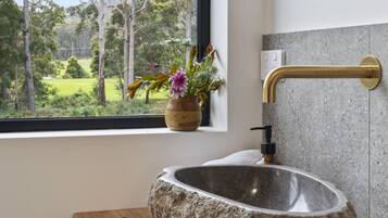 A stone sink with a brass faucet, a window with a view of a forest, and a vase with flowers on the windowsill.