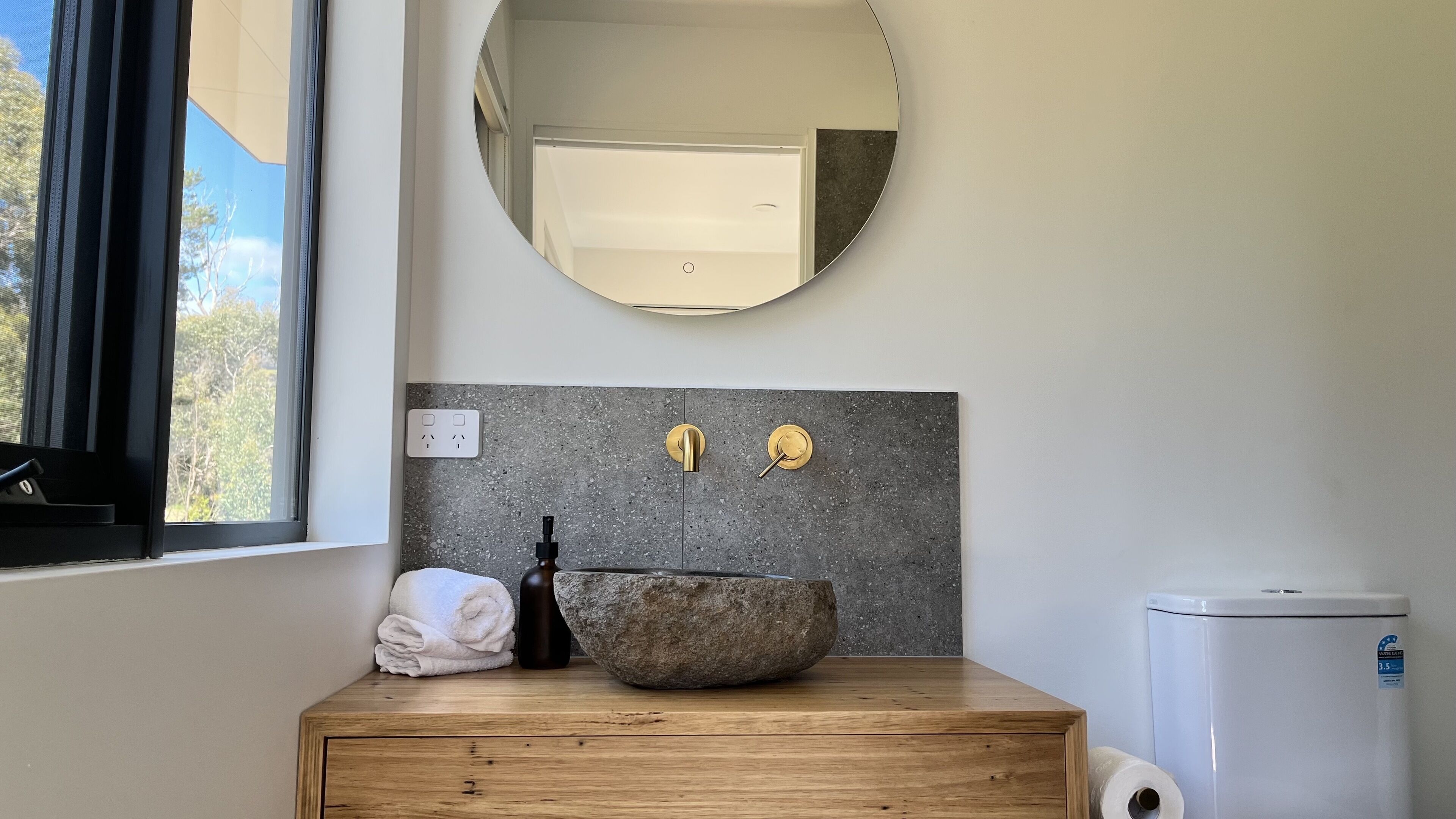 Een badkamer met een houten wastafelmeubel, een ronde spiegel, een stenen wastafel en een witte prullenbak.