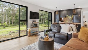 Een moderne woonkamer met een groot raam, een zwarte leren bank, een houten salontafel en een keuken met donkere kastjes en een roestvrijstalen koelkast.