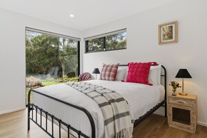 Quarto moderno com uma cama grande, duas janelas, mesa de cabeceira com luminária e vista para o verde lá fora.