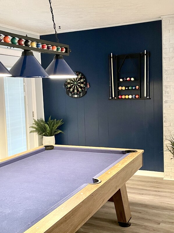 A pool table with a dartboard and a rack of billiard balls against a dark blue wall.