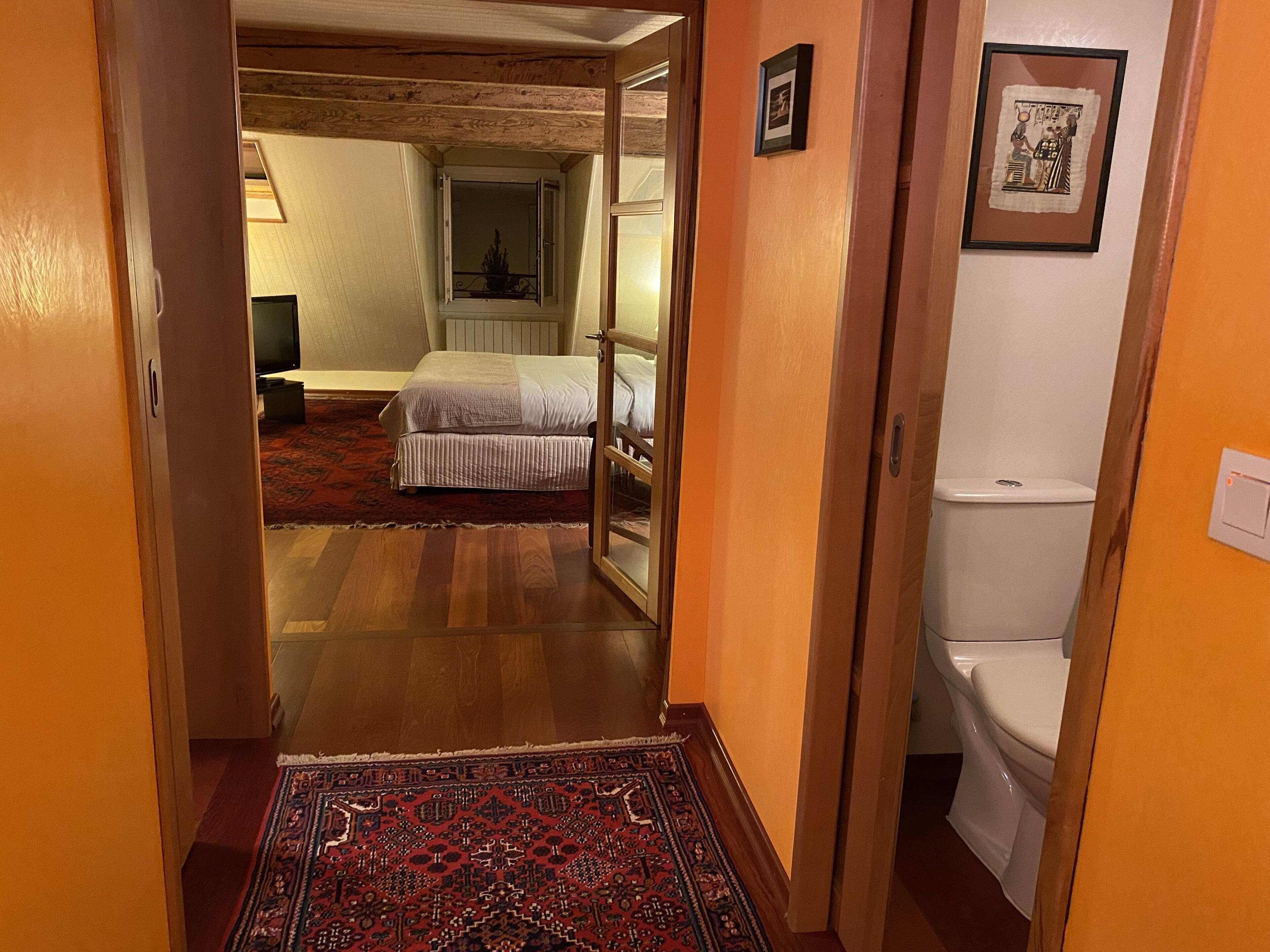Ein Flur, der in ein Schlafzimmer mit Bett, kleinem Tisch und Stuhl führt. Ein Badezimmer mit Toilette und Dusche ist ebenfalls zu sehen.