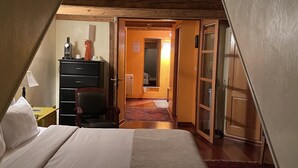 Een slaapkamer met een groot bed, houten balken, een bureau met lamp en uitzicht op een gang en badkamer.