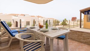 A terrace with white furniture, a table set with plates, glasses, and a bottle, and a view of buildings and palm trees. - 2 Bed Hotel Apt, Sleeps 4/6, Costa Adeje, 5 minutes walk to Beach, Shops & Bars (Costa Adeje)