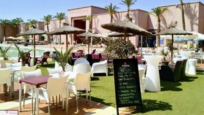 Zona pranzo all'aperto con sedie e tavoli bianchi, una piscina sullo sfondo e palme.