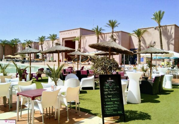 An outdoor dining area with white chairs and tables, a pool in the background, and palm trees.