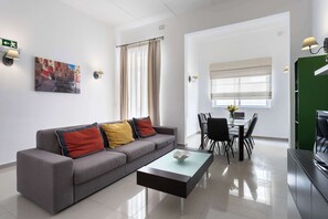 Een moderne woonkamer met een grijze bank, een glazen salontafel, een eethoek met een tafel en stoelen, en een groene deur.