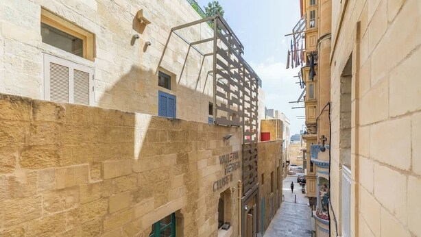 Une ruelle étroite entre des immeubles aux façades en pierre, une structure métallique et un ciel dégagé.